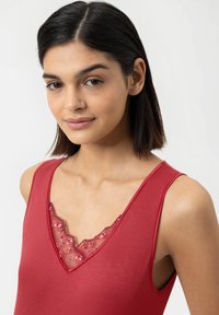 Young woman with short dark hair wearing a red sleeveless top with floral lace detail at the neckline against a plain background.