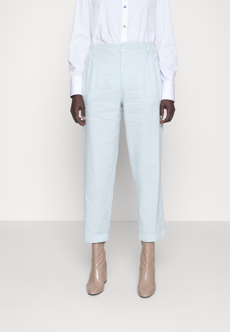 Personne portant un pantalon taille haute bleu clair, une chemise blanche à boutons et des bottines beiges, debout devant un fond clair uni.