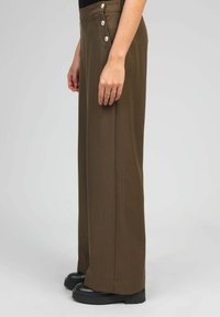 Pantalons beige à taille haute, larges, avec des boutons décoratifs sur le côté et une texture lisse. Portés avec des chaussures à plateforme noires.