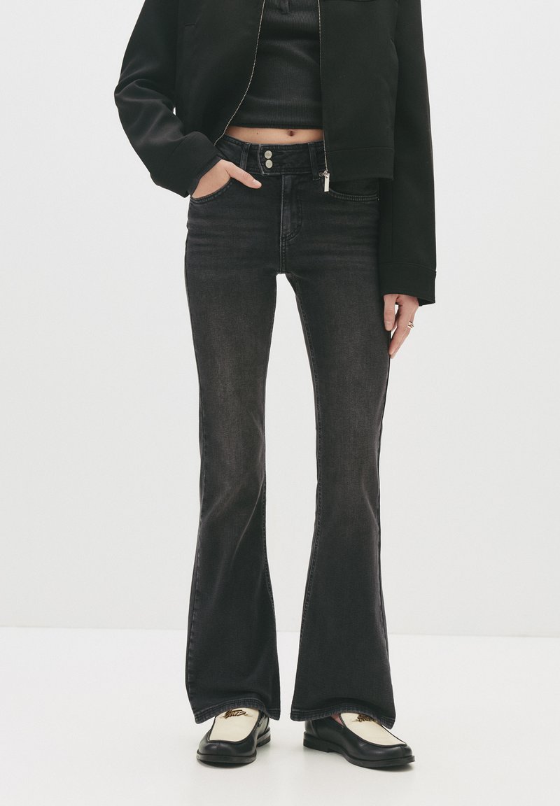 Person som bär svarta utsvängda jeans, svart kort jacka, svart kort topp och svart-vita loafers, står mot en vit bakgrund.