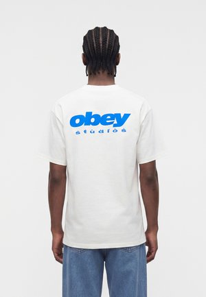 Hombre con trenzas, lleva una camiseta blanca con un gran texto azul "obey studios" en la parte trasera, combinada con vaqueros azul claro, de espaldas.