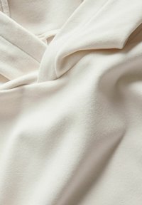 Svetlo bež tkanina z mehko, rebrasto teksturo. Material se nežno zloži in razkriva prekrivne detajle blizu izreza na vratnem predelu.