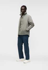 Veste matelassée gris clair avec col montant, fermeture à boutons-pression ; portée avec un pantalon bleu foncé et des baskets blanches.