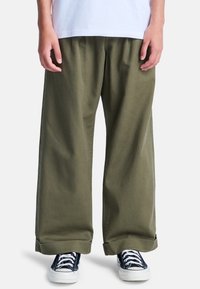 Pantaloni larghi in cotone verde oliva con una texture morbida e risvolti, abbinati a sneakers blu navy con suole e lacci bianchi.