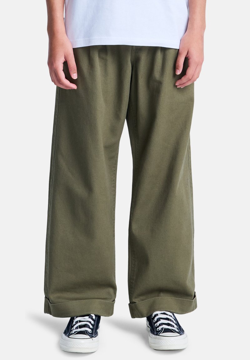 Pantaloni larghi in cotone verde oliva con una texture morbida e risvolti, abbinati a sneakers blu navy con suole e lacci bianchi.