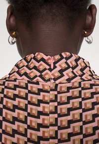 Cheveux courts et robe à motifs géométriques dans des tons de rose, noir et marron avec une fermeture éclair au dos, portée par une personne à la peau foncée et portant des boucles d'oreilles transparentes.