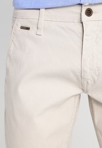 Chinos in cotone beige con una texture liscia, dotati di chiusura a bottone e di un piccolo logo metallico sulla tasca frontale.