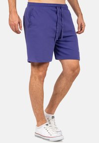 Pantaloni corti in cotone viola con vita elasticizzata, cordoncino e due tasche laterali. Il design presenta un logo sulla parte inferiore della gamba.