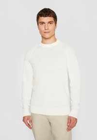 Weißer Strickpullover mit rundem Hals, gerippten Bündchen und Saum, getragen über hellbeigen Hosen. Verfügt über ein strukturiertes Muster.