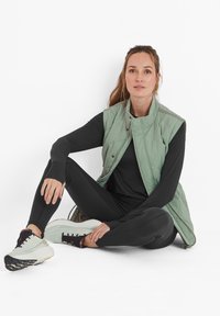 Light green quilted vest, black long-sleeve top and leggings, pale sneakers with dark accents, sitting posture, neutral background.