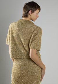 Guldglinsende cropped top med korte ærmer og en blød, struktureret overflade. Har en bred krave og en tætsiddende silhuet.