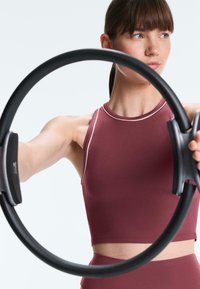 Ženska v bordo športni obleki drži črni Pilates obroč z obema rokama in izteguje roke naprej na belem ozadju.