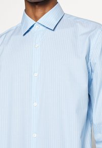Uomo che indossa una camicia button-up a righe verticali bianco e azzurro chiaro con colletto classico, bottoni allacciati.