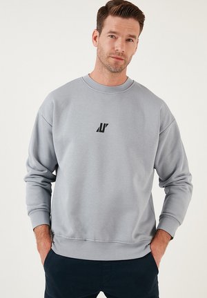 Light gray sweatshirt featuring a round neckline, ribbed cuffs, and hem, with a small black logo on the chest. Soft fabric texture.