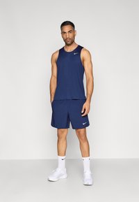 Σετ αμάνικη μπλούζα και σορτς σε ναυτικό μπλε, κατασκευασμένο από ελαφρύ ύφασμα. Διαθέτει λευκό λογότυπο Nike στο στήθος και στα σορτς. Φορεμένο με λευκά αθλητικά παπούτσια.