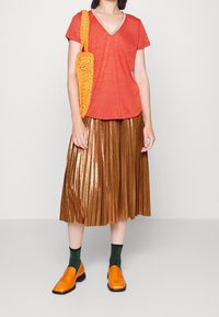 Rostfärgad v-ringad t-shirt, metallisk bronserad plisserad kjol, orange stickad väska, orange loafers och gröna texturerade strumpor.