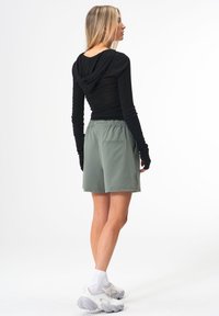 Schwarzes Langarmshirt mit Kapuze, kombiniert mit hellgrünen Shorts mit elastischem Bund und einer Gesäßtasche. Weiße Sneakers mit grauen Akzenten.