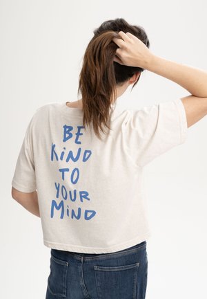 Vrouw met bruin haar die een paardenstaart vasthoudt, gekleed in een wit shirt met blauwe tekst waarop staat "WEES VRIENDELIJK VOOR JE GEEST" en een blauwe spijkerbroek, met de rug naar de camera gekeerd.