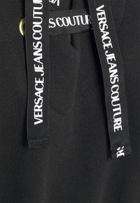 Tissu noir avec cordons noirs présentant le texte blanc "Versace Jeans Couture" imprimé de façon répétée.