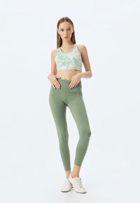 Groene sportieve crop top met een tie-dye patroon, gecombineerd met hoge, effen groene legging en witte sportschoenen, staande met de handen op de heupen.