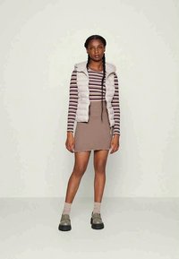 Colete acolchoado em cinza claro, camisa de manga longa listrada em roxo e branco, saia canelada castanha, sapatos de plataforma cinzas e meias até ao joelho.