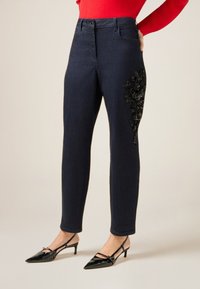 Jeans de mezclilla azul oscuro con cintura alta, que presentan un adorno de lentejuelas florales negras en el lado izquierdo. Combinados con tacones de punta afilada negros.