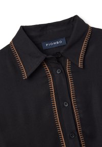 Camisa negra con botonadura en la parte delantera, costuras marrones contrastantes a lo largo del cuello y la tapeta, con una etiqueta de marca.