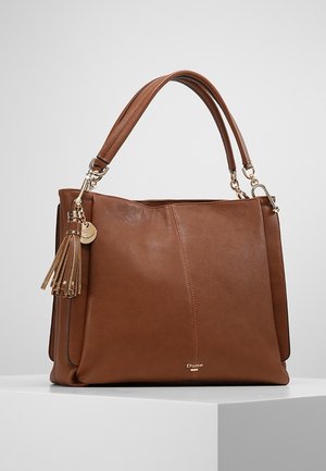 Brązowa skórzana torebka z podwójnymi uchwytami, złotym okuciem, ozdobą z chwostem i logo "Dune" z przodu, umieszczona na białym postumencie.