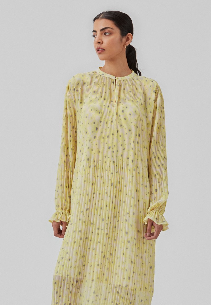 Robe jaune plissée avec des motifs en points gris, manches longues, poignets volantés et un décolleté en œil de clé. Tissu léger et aéré.