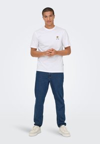 White cotton t-shirt with short sleeves and a small graphic on the left chest, paired with blue denim jeans and light-colored sneakers.
