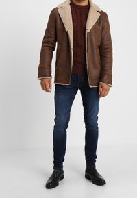 Brązowa skórzana kurtka z kożuchem i kremowym kołnierzem, w zestawieniu z bordowym swetrem w warkoczowy splot, ciemnoniebieskimi jeansami i czarnymi botkami za kostkę.