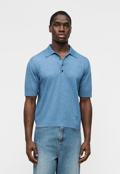Homme portant un polo à manches courtes bleu avec des boutons et un jean bleu clair, debout devant un fond uni.