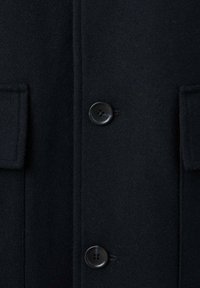 Abrigo de lana negro con dos botones negros y dos bolsillos con solapa en la parte delantera, mostrando un primer plano de la textura de la tela y los detalles de costura.