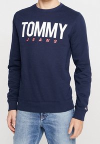 Námořnicky modrý sweatshirt z bavlny s kulatým výstřihem. Na přední straně se nachází velké bílé logo "TOMMY" a červený text "JEANS". Hřejivé elastické manžety a lem.