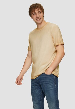 Korte mouwen, ronde hals t-shirt gemaakt van lichte stof in een verloop van goudkleur, gecombineerd met donkerblauwe jeans, handen in de zakken.
