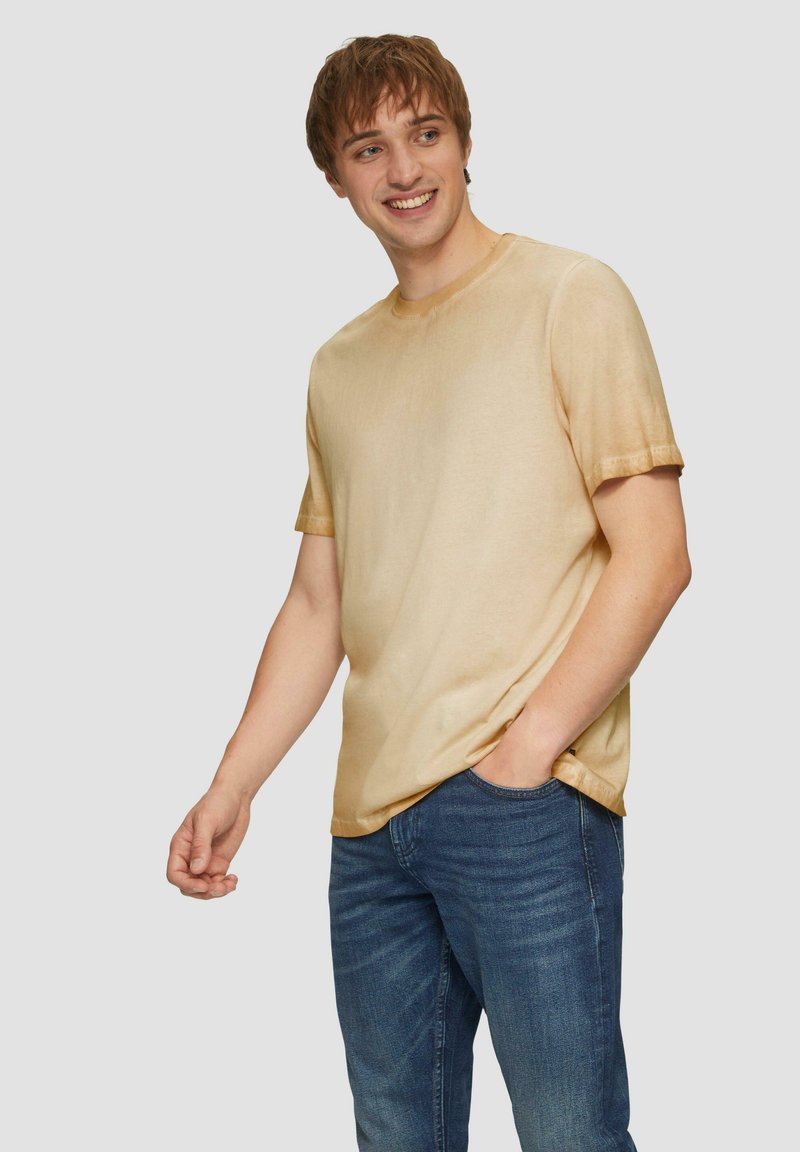 Kurzarm-T-Shirt mit Rundhalsausschnitt aus leichtem Stoff in einem goldfarbenen Farbverlauf, kombiniert mit dunkelblauen Jeans, Hände in den Taschen.