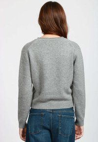 Donna con lunghi capelli castani, che indossa un maglione grigio e jeans blu, vista di spalle su uno sfondo chiaro e uniforme.