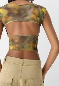 Genomskinlig, kort topp med en gradientmönster i grönt och gult, med en öppen ryggdesign och kontrasterande kant. Bärs med beige byxor.