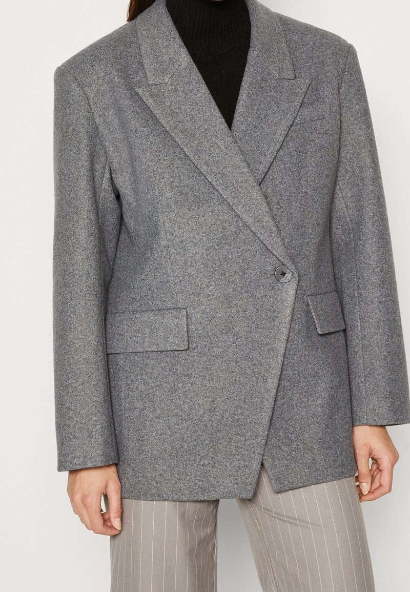 Personne portant un blazer en laine gris à un seul bouton sur un col roulé noir, associé à un pantalon beige à rayures fines.