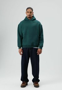 Groene hoodie met capuchon en zak, losse pasvorm. Gecombineerd met donkerblauwe wijde pijpen jeans en bruine veterschoenen. Effen achtergrond.
