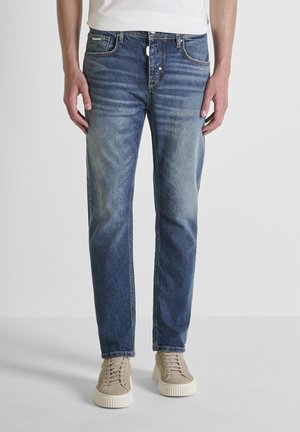 Modello maschile che indossa jeans blu slavati a gamba dritta e sneakers beige, in piedi contro uno sfondo chiaro uniforme.