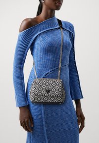Woman in a blue ribbed off-shoulder dress wearing a black and white patterned Guess crossbody bag with gold chain strap.