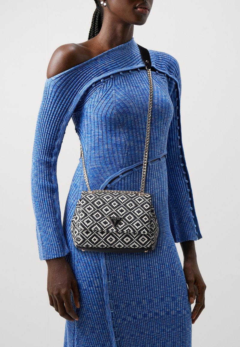 Woman in a blue ribbed off-shoulder dress wearing a black and white patterned Guess crossbody bag with gold chain strap.