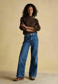 Brauner Strickpullover mit Seitenschaltern, kombiniert mit hochgeschnittenen blauen Denim-Baggy-Jeans. Die Schuhe haben eine Mischung aus Braun, Rot und Grau.