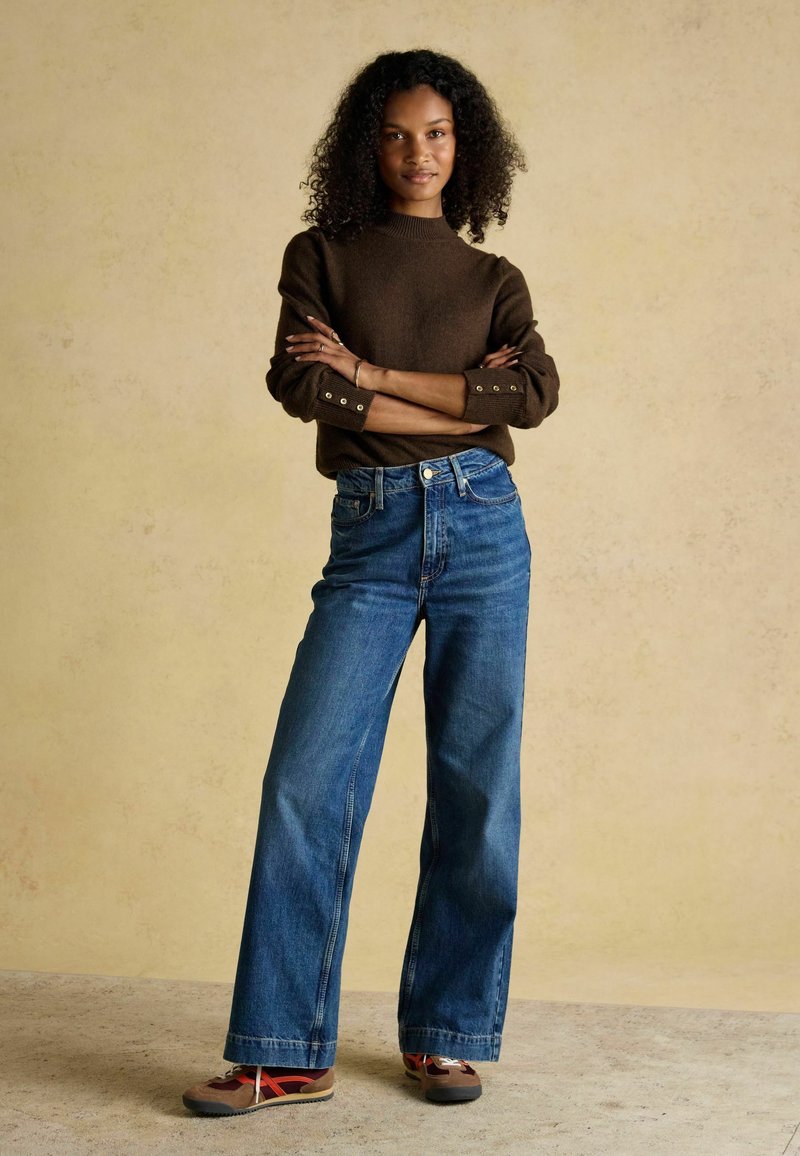 Brauner Strickpullover mit Seitenschaltern, kombiniert mit hochgeschnittenen blauen Denim-Baggy-Jeans. Die Schuhe haben eine Mischung aus Braun, Rot und Grau.