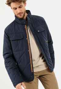 Marineblauer gesteppter Mantel mit Stehkragen, brauem Reißverschluss und zwei Brusttaschen. Weiche Textur mit brauner Innenauskleidung.