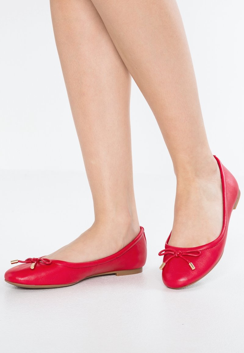 Ballerines en cuir rouge avec un bout arrondi, dotées d'un détail nœud avec des accents dorés et d'un petit talon plat. Texture lisse.