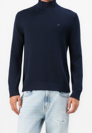 Mężczyzna ubrany w granatowy golf i jasnoniebieskie, przetarte dżinsy, stojący na tle białego tle.