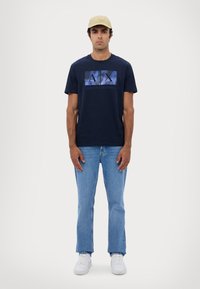 T-shirt in cotone blu navy con logo "AX" e testo "ARMANI EXCHANGE"; abbinata a jeans azzurri e sneaker bianche, completata con un cappellino beige.