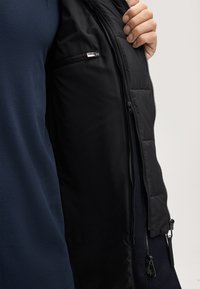 Giacca nera imbottita con tasca interna con zip, dal tessuto liscio e dalla vestibilità aderente, indossata sopra una camicia blu navy.